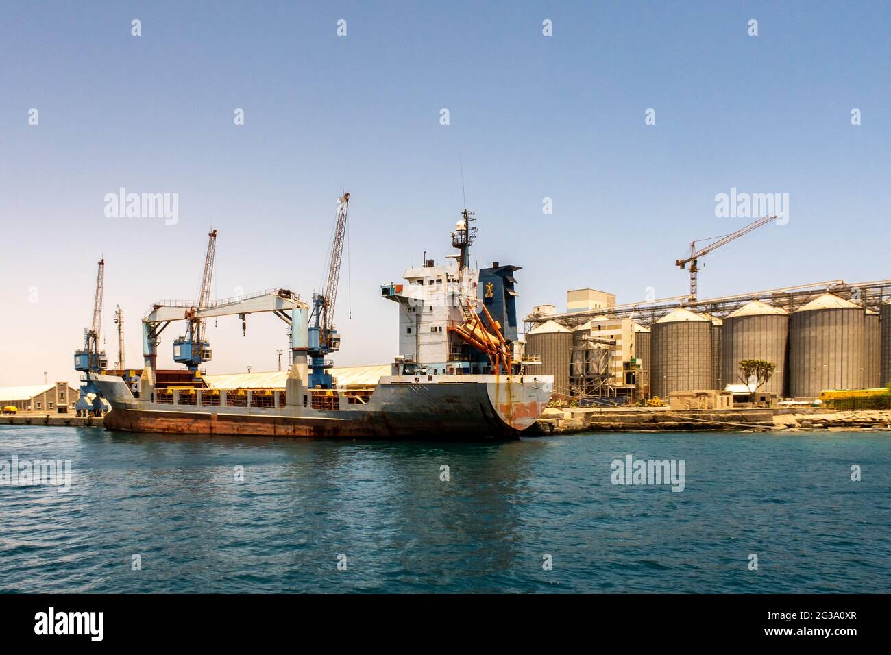 Large container ship being loaded with cargo in port in Port Sudan. Huge port cranes and row od grain silos in the background. Stock Photo