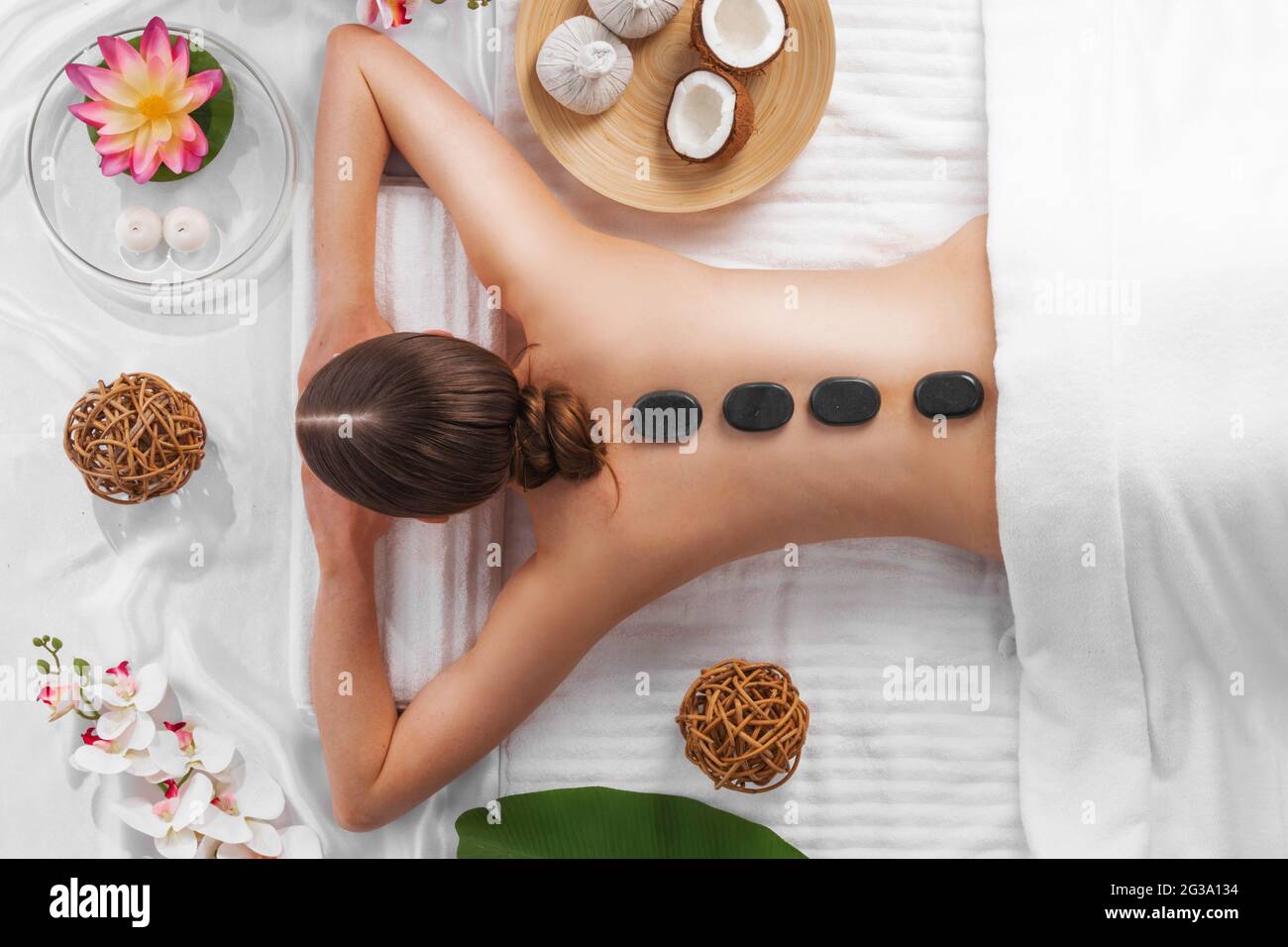Spa stone massage, beautiful woman getting hot stones massage, beauty treatments concept. Top view. Orchid and lotus flowers coconut and herb pouches Stock Photo