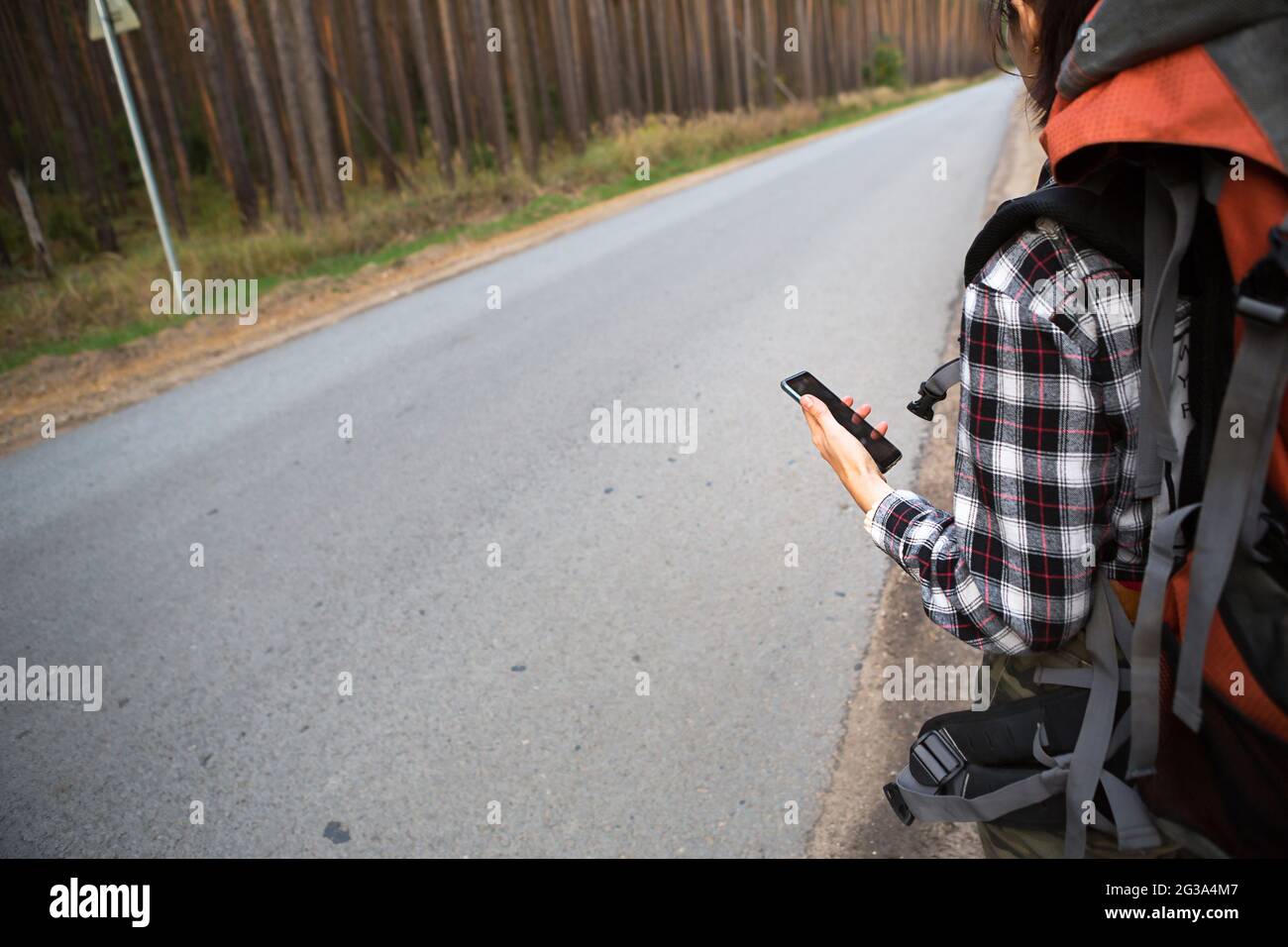 Female tourist in a check shirt with an orange large backpack near a highway in the woods with a smartphone in hand. Navigation, satellite maps, commu Stock Photo