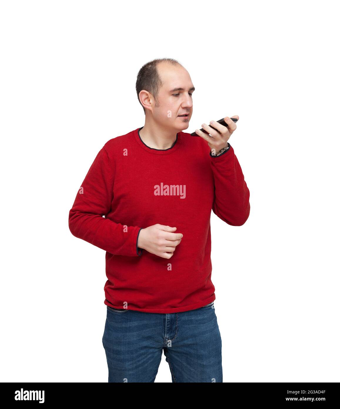 A bald young man is recording an audio note with his mobile phone. The  person is dressed in a red sweater, jeans and wears a watch. The background  is Stock Photo -