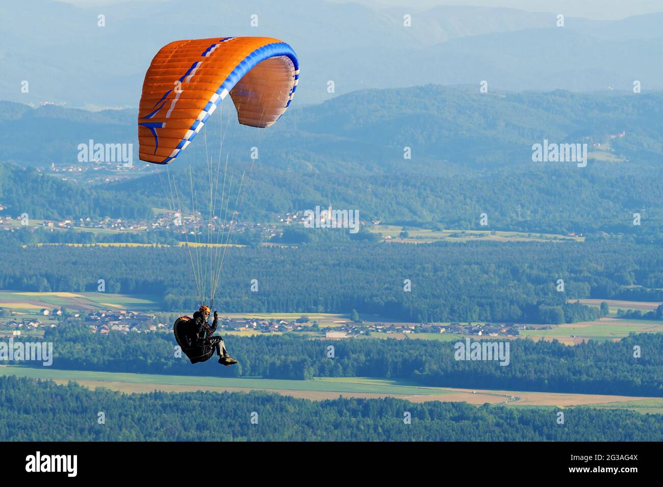 Paraglider with orange parachute flying over countryside before sunset. Adrenaline, extreme sports and relaxation concepts. Stock Photo