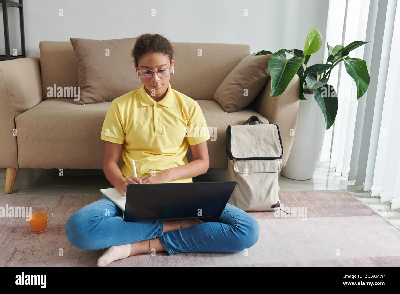 Serious mixed race girl in glasses and ear buds sitting with crossed legs on floor and making notes in workbook while learning lesson online Stock Photo