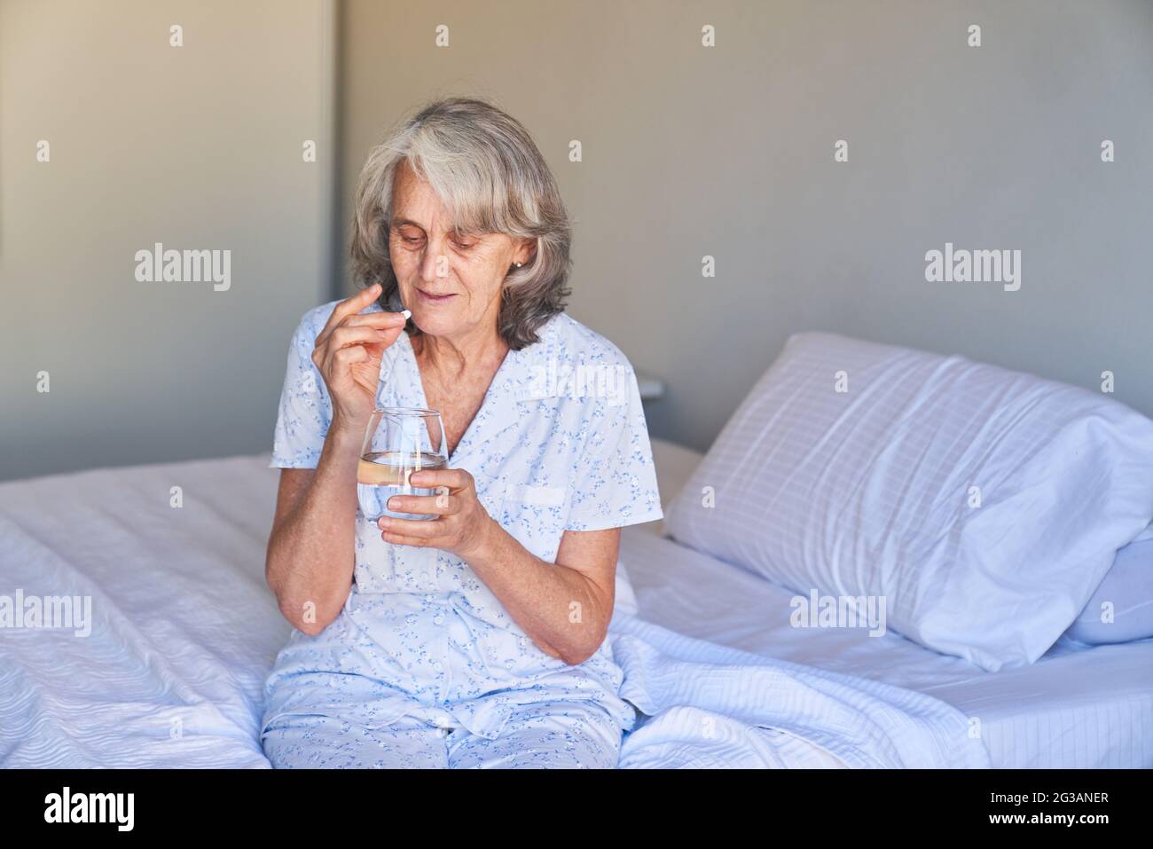 Sick elderly woman sits on the bed in the hospital and takes a tablet with water Stock Photo