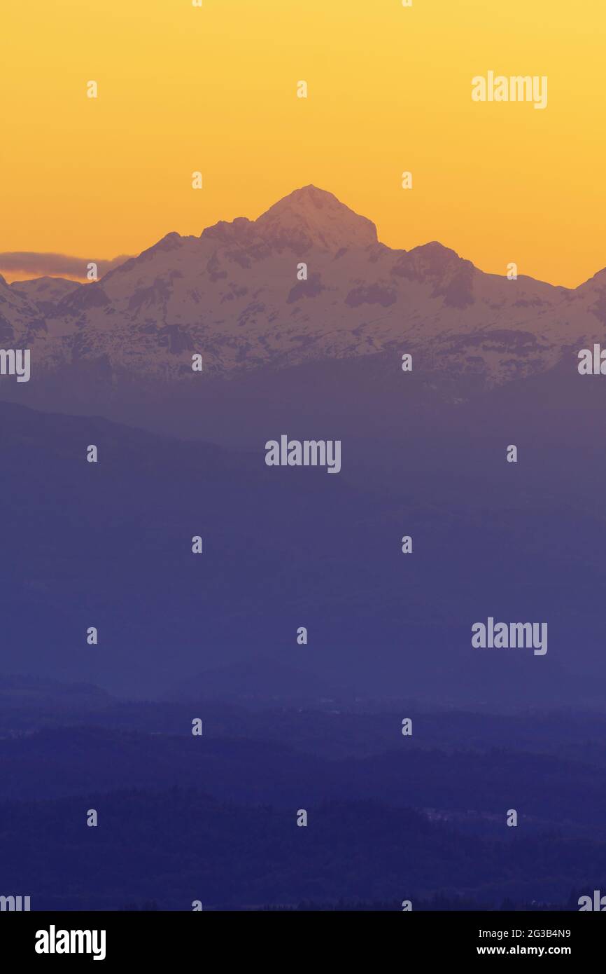 Triglav, the highest alpine peak in Slovenia during twilight time with intensely orange sky in background and layers of hills in foreground. Beautiful Stock Photo