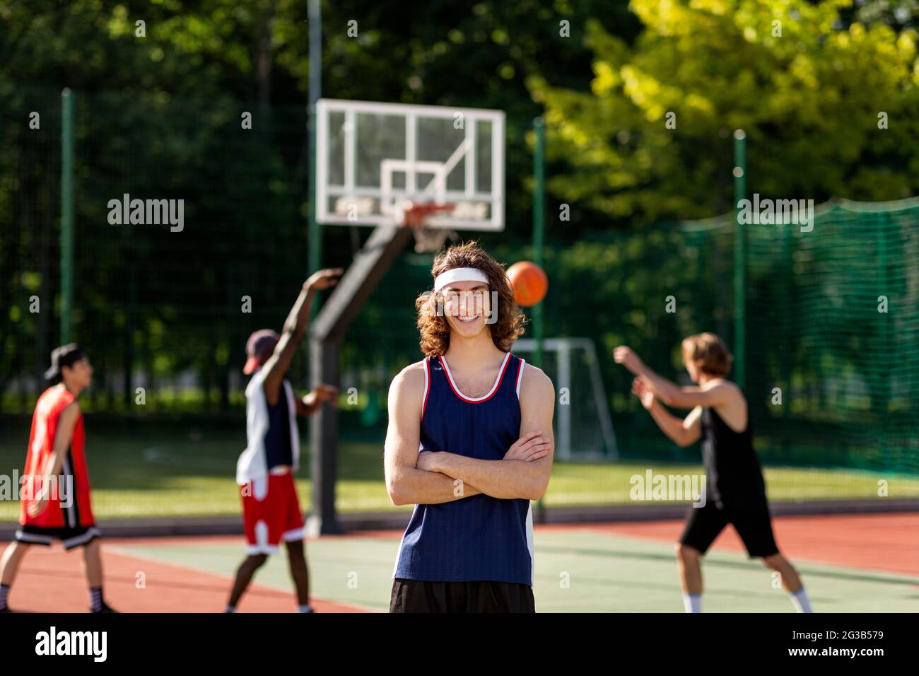 Happy Caucasian sportsman posing with crossed arms and smiling at camera,  his diverse team playing basketball at outdoor arena, copy space. Summertime  Stock Photo - Alamy