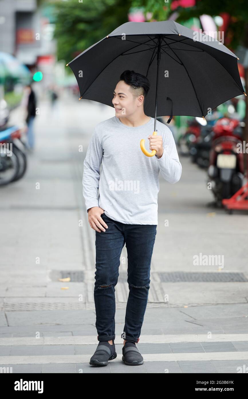 Cheerful young Asian guy in casual wear holding umbrella and walking in rainy weather over city street with scooters Stock Photo