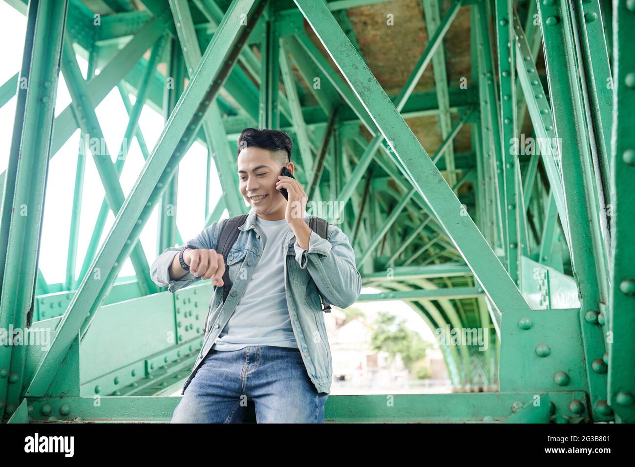 Smiling young Asian man with satchel sitting on metal girder and checking time while calling by phone under bridge Stock Photo