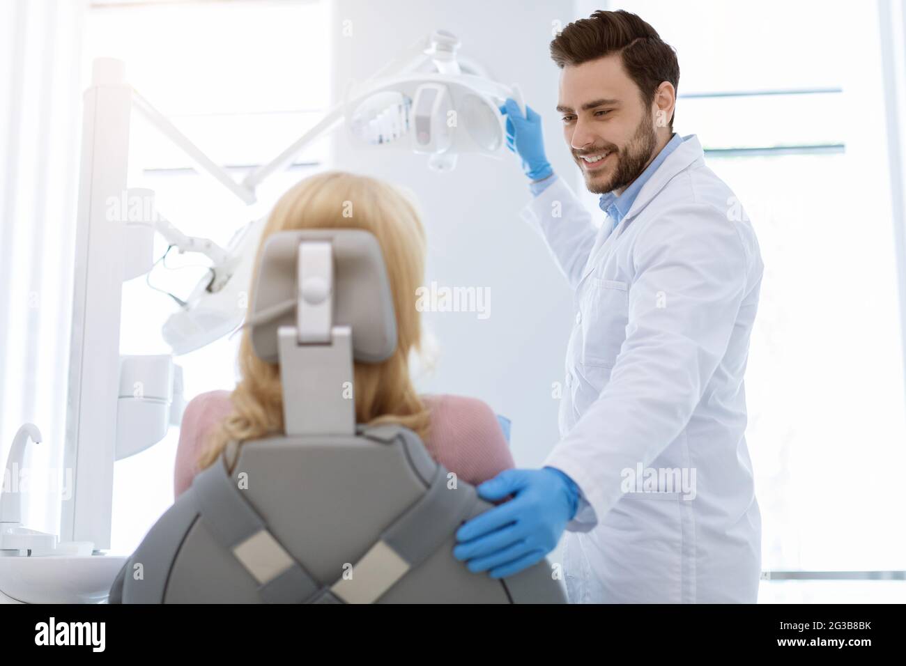 Young handsome bearded man dentist doctor greeting and comforting female patient sitting in dentist chair, turning on lamp adn smiling, wearing rubber Stock Photo