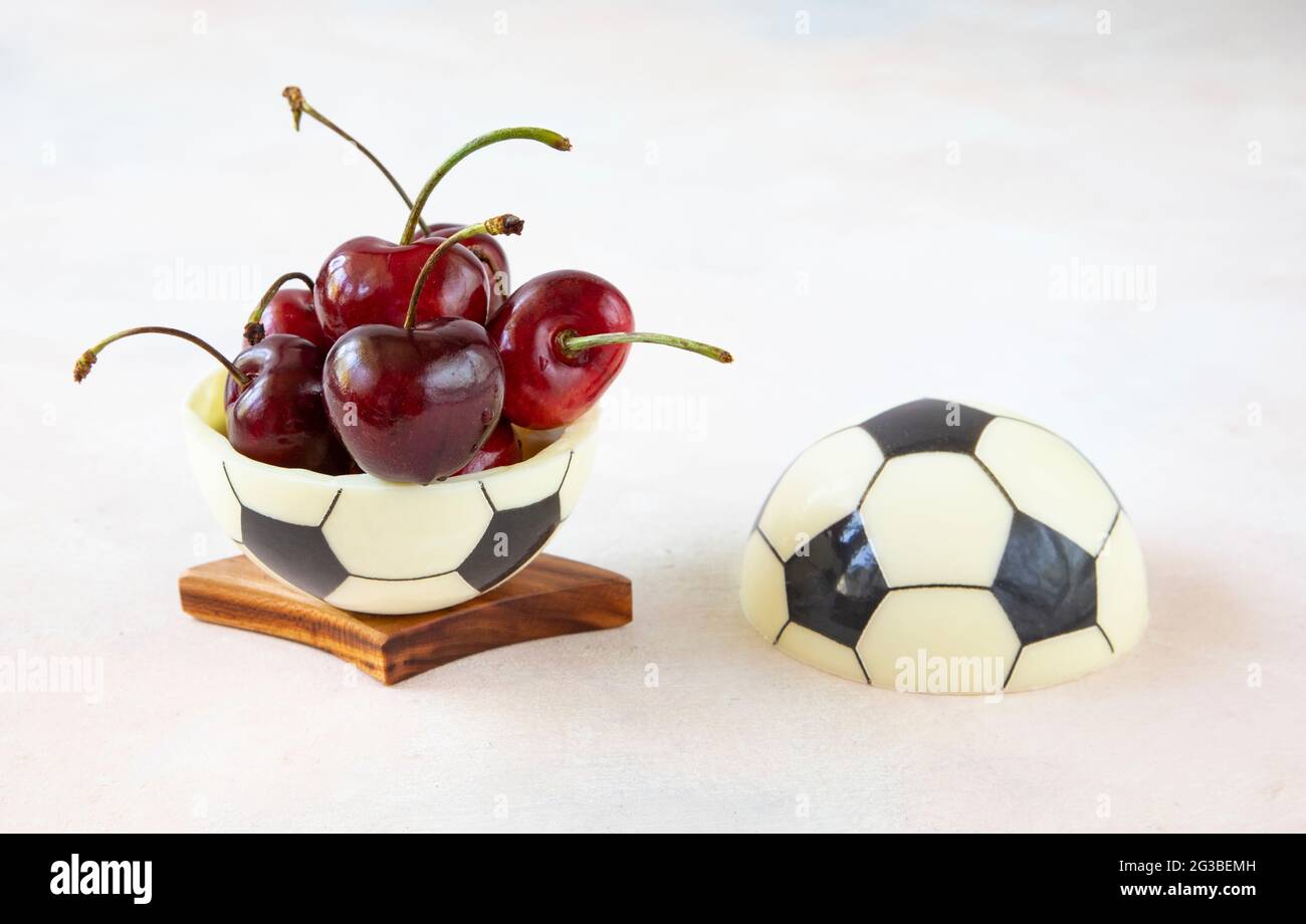 A sweet dessert for a fan of european football - a chocolate black and white ball made of two halves with ripe red cherries with sprigs on the table Stock Photo