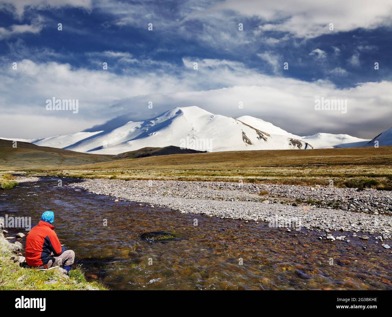 Man sitting alone on the river bank against snowy mountain and blue sky background, travel concept Stock Photo