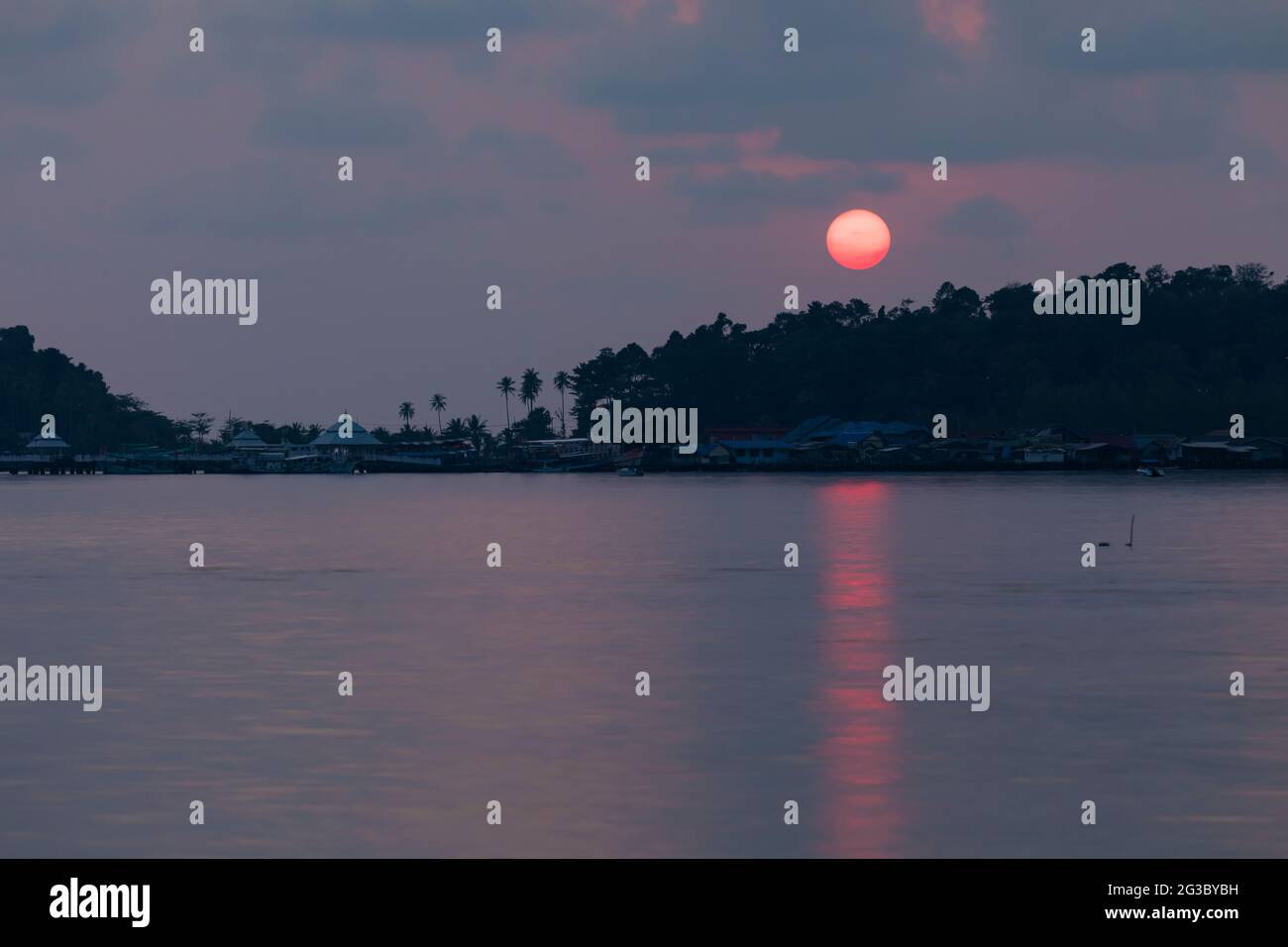 A huge sun, tinted red, sets behind the rainforest that surrounds the small town of Bang Bao, on the island of Koh Chang, Gulf of Thailand Stock Photo