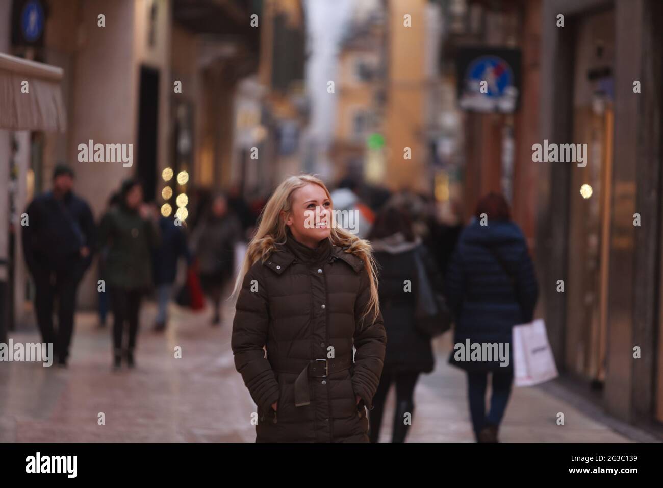 Happy natural young woman strolling down a busy urban pedestrian street in a warm winter coat looking up to the side with a smile with lateral copyspa Stock Photo