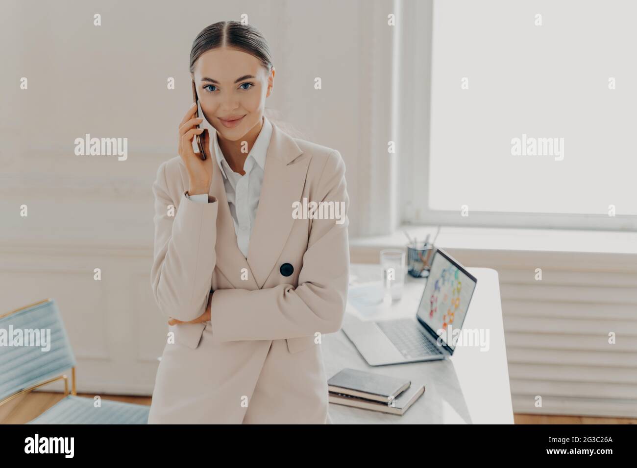 Positive young female entrepreneur talking on phone and smiling at camera Stock Photo