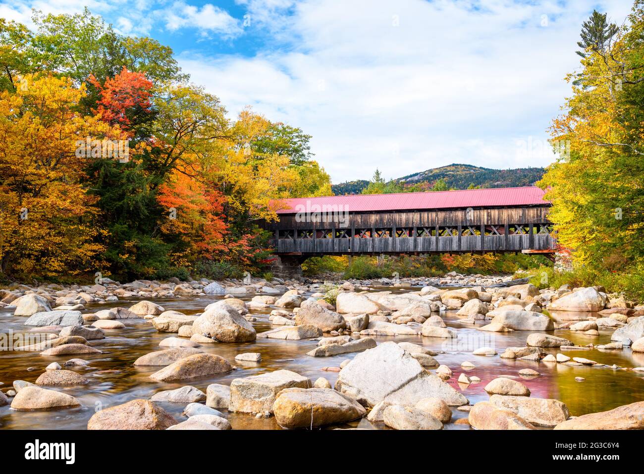 Old covered bridge spanning a mountain river with forested banks on a partly cloudy autumn day. Stunning autumn colours. Stock Photo