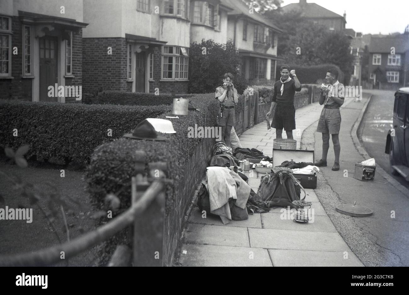 1951, historical, we made it!...outside in a suburban street, by a car of the era, two scout masters and one young scout, stand by their baggage and camping gear, pots and pans which are lying on the pavement. One of the men raise's a hand pump in salute to the successful end of their scout camp trip, England, UK. Stock Photo