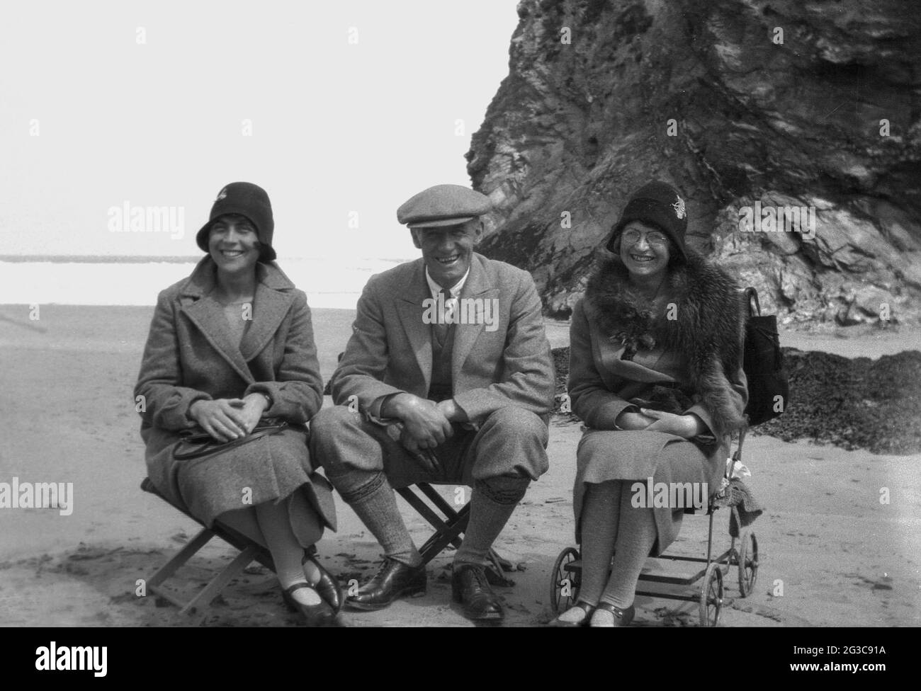 1930s, historical, all smiles as an English gentleman and two ladies sit beside each other on small stools on a beach in England, UK, wearing the formal clothing of the era for a photo. The two women are in coats and the hats of the day, while the man is wearing plus fours, jacket & tie and a wide cloth cap. Stock Photo