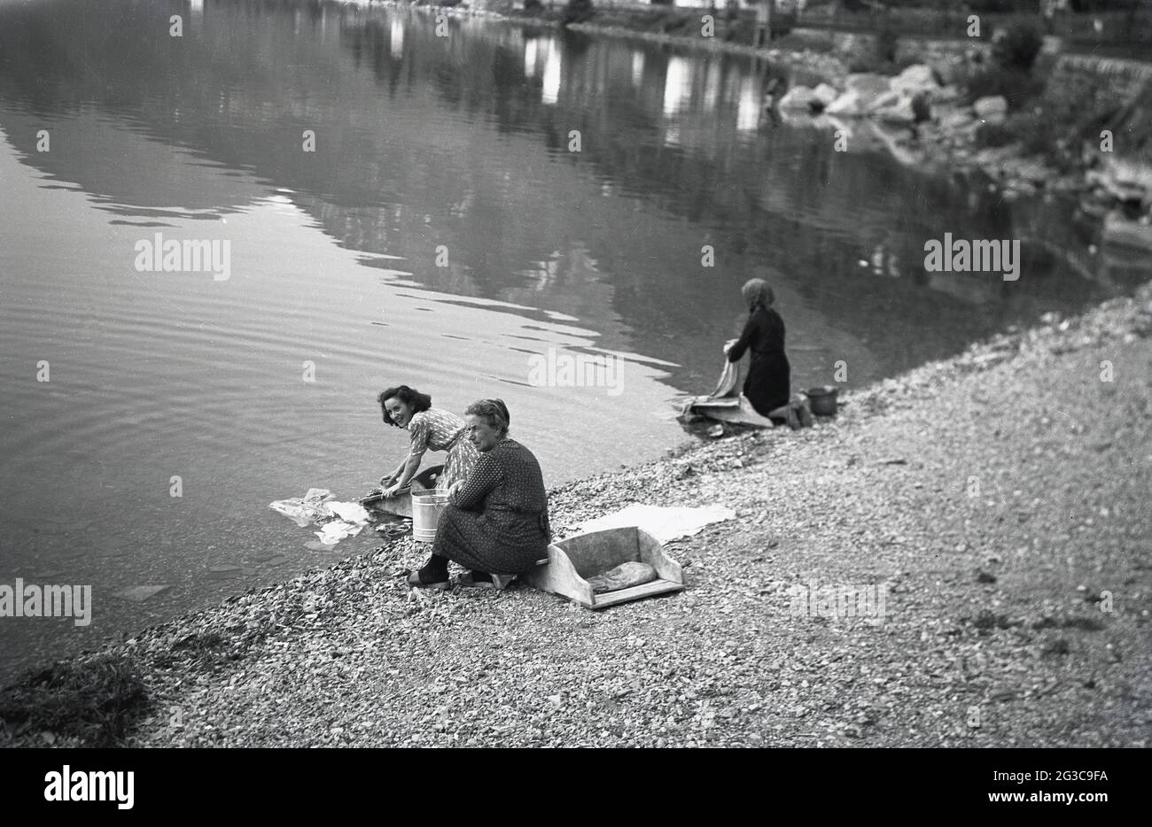 1950, two women on a shore by a lake hand washing garments using a form of washboard, a wooden washing box and board, France. This three-sided wooden box or caisse had a back half with a place to kneel on and front half (or washboard) where the clothes would be scrubbed or washed with the water. The woman knelt in the box and her skirt stayed dry. For more comfort, it would be padded with straw or a cushion as seen here. In some areas of the world, doing the laundry by a local river or lake, a tiring yet effective method, is still practised today. Stock Photo