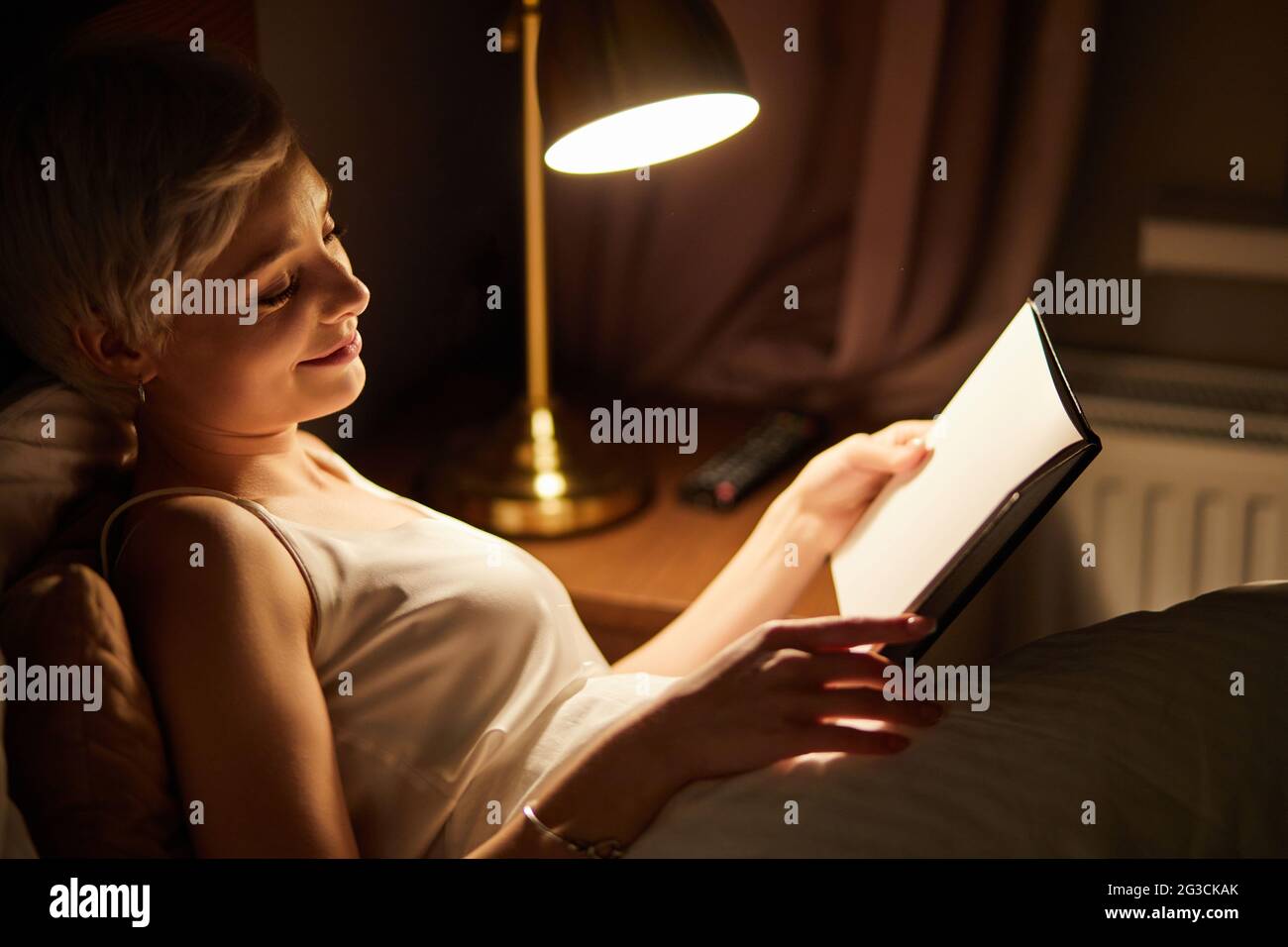 Beautiful female in pajamas lying on bed reading book, alone at night. Caucasian short haired lady in bedroom, charming cute woman in room lighted by Stock Photo