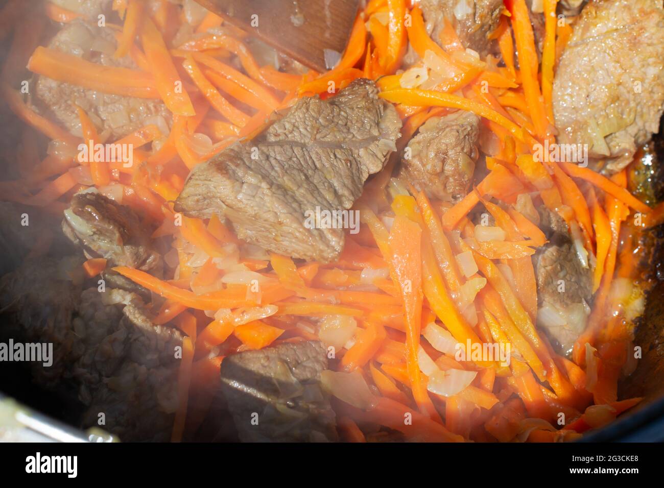 Cooking traditional oriental pilaf in a cauldron over a fire. Mix large pieces of grilled meat and finely chopped vegetables and simmer for a while. C Stock Photo