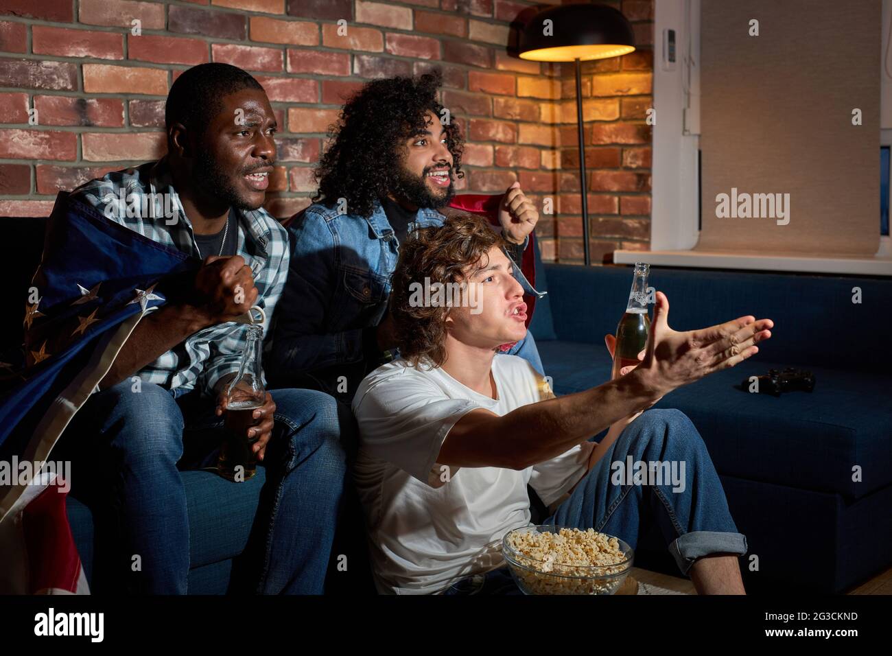 Group Of Diverse Men Sitting On Sofa Watching Sport Championship Together Emotionally Reacting On Game Process, Gesturing, Drinking Beer, At Home Stock Photo