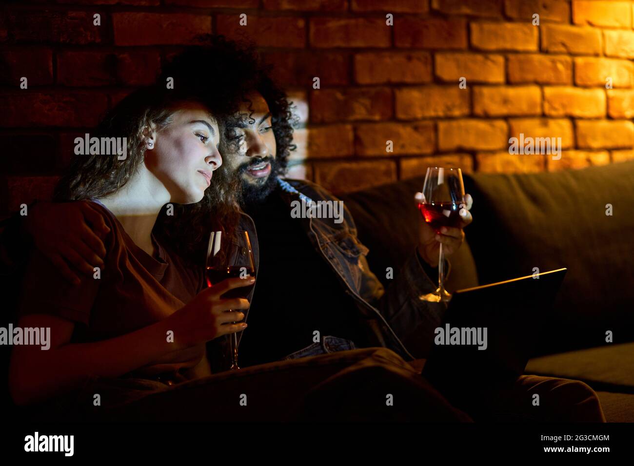 Diverse married couple drinking red wine while watching films, movies, comedy. Beautiful woman love spending time with boyfriend at home Stock Photo