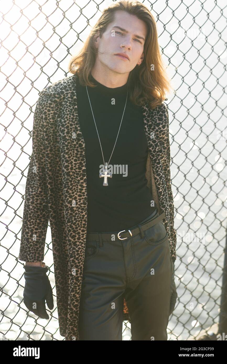 A young male model posing outdoor in a long winter coat, posing againt a chain link fence. He has long brown hair, wears a black leather glove. Stock Photo