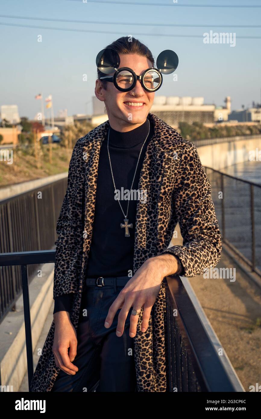 A young male model posing outdoor in a long winter coat, he wears a fun mickey mouse ears sunglasses and a cross necklace. A fun fashion portrait. Stock Photo