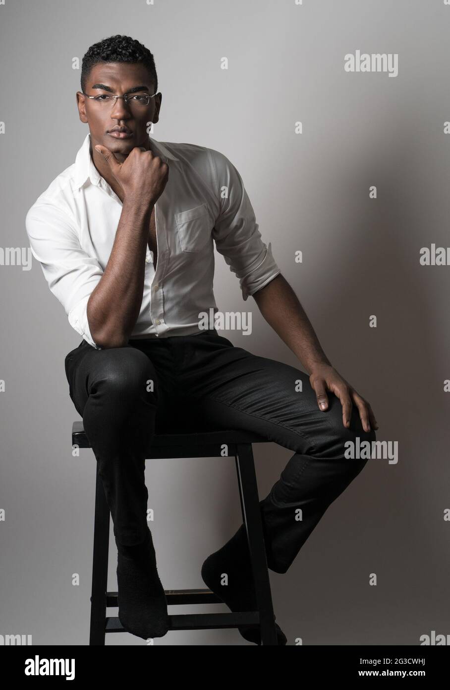 A young black male wearing glasses, a fashion model, sitting on a wooden stool posing with his hand to his chin, a serious portrait. Stock Photo
