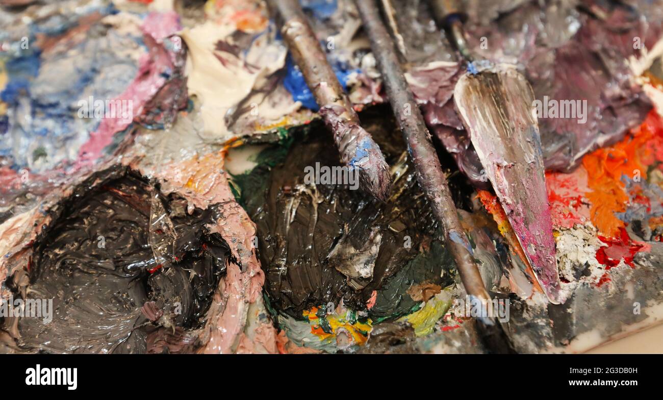 An artist's or painters palette covered with mixed thick rich oil colour. Palette knife and brushes laying diagonally across the mostly purple, blue a Stock Photo