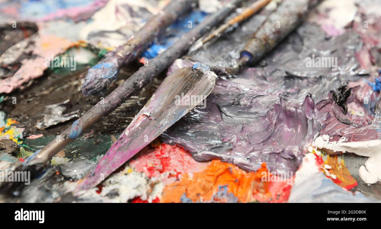 An artist's or painters palette covered with mixed thick rich oil colour. Palette knife and brushes laying diagonally across the mostly purple, blue a Stock Photo