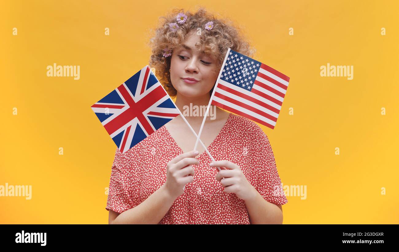 Beautiful young girl holding flags of USA and UK in her hands. Girl isolated in yellow background studio. Dressed in pink shirt and wearing flowers in her hair. Concept of English Speaking countries.  Stock Photo