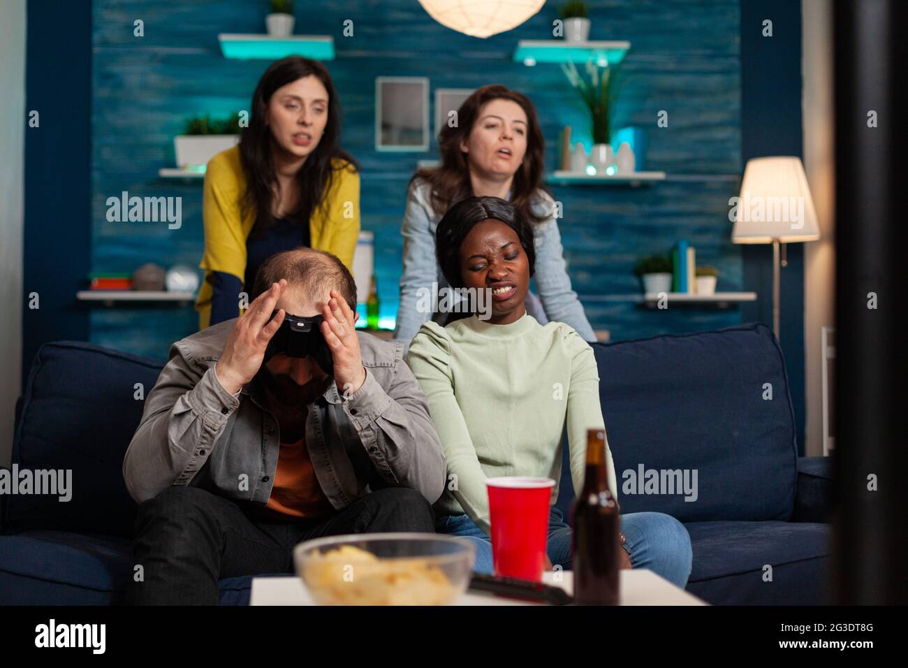 Angry man and multi ethnic friends upset after losing game competition, bonding and sitting on couch after drinking beer. Mixed race group of people hanging out together having fun late at night in living room Stock Photo