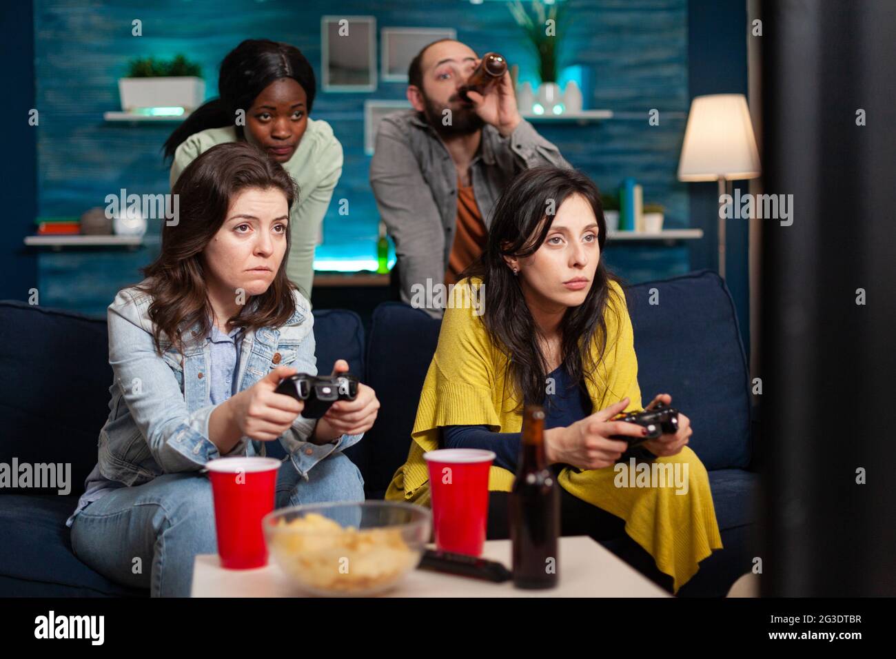 Multi ethinc friends cheerful group of people relaxing on console games with controller. Group of mixed race friends playing games while sitting on sofa in living room late at night. Stock Photo