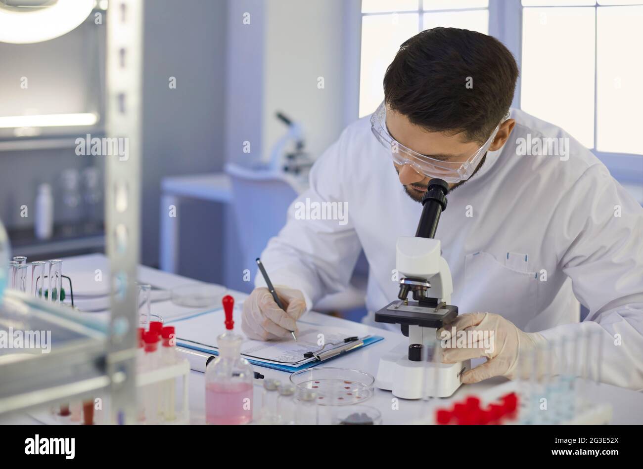 Scientist taking notes sitting at table with microscope in microbiology laboratory Stock Photo