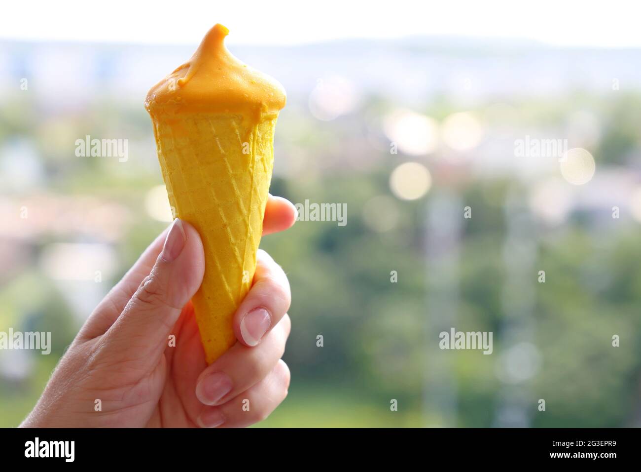 Ice cream in waffle cone in female hand on summer city background. Cool dessert with mango juice in hot weather Stock Photo