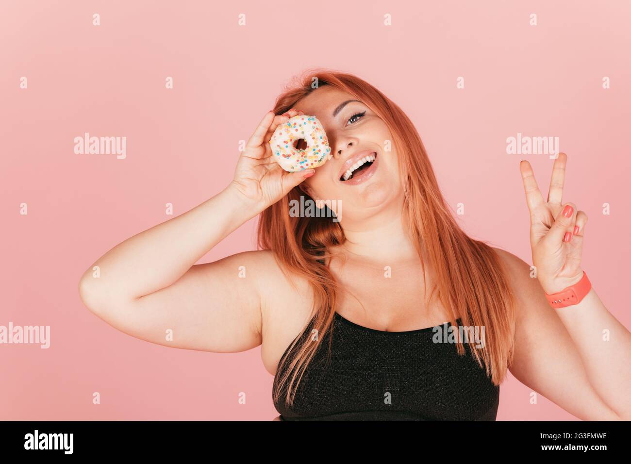 Joyful oversized young woman laughs holding dessert donut near her eye and shows peace gesture while standing on pink background. Stock Photo