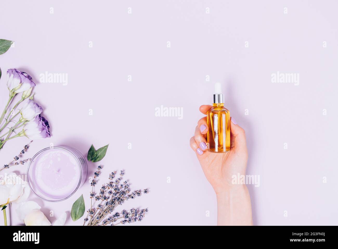 Female hand holding bottle of cosmetic oil next to scented candle, lavender, white peony and lisianthus flowers on light purple background, flat lay. Stock Photo