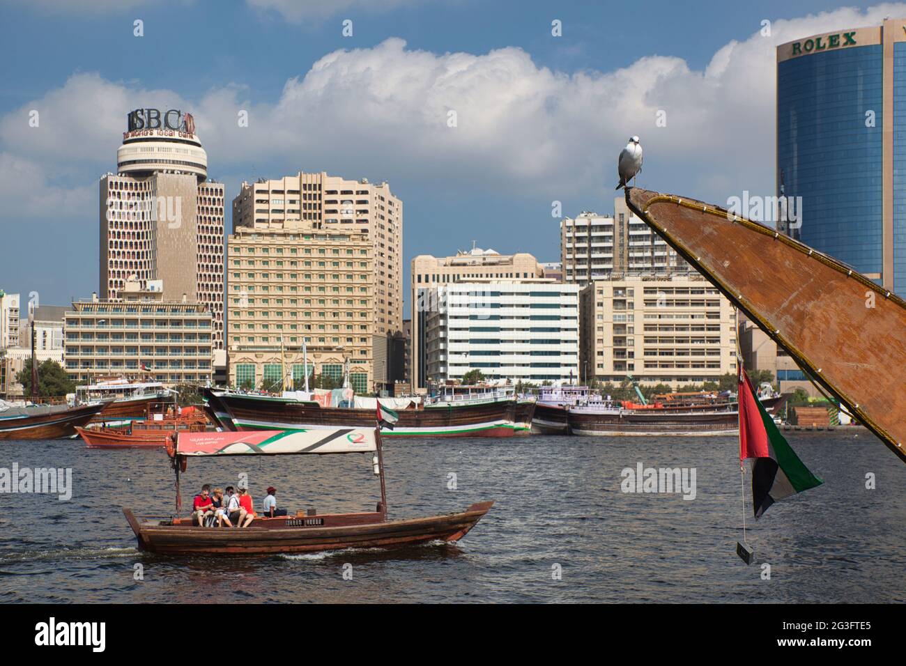 Dubai Creek, the river that flows through Dubai with a water taxi sailing and prow of boat with a seagulsl perched on tip and city high risers beyond Stock Photo