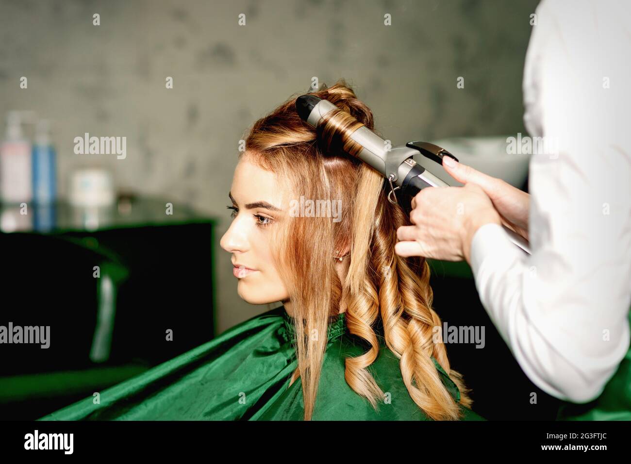 Hairdresser using curling tongs curls long brown hair on the young
