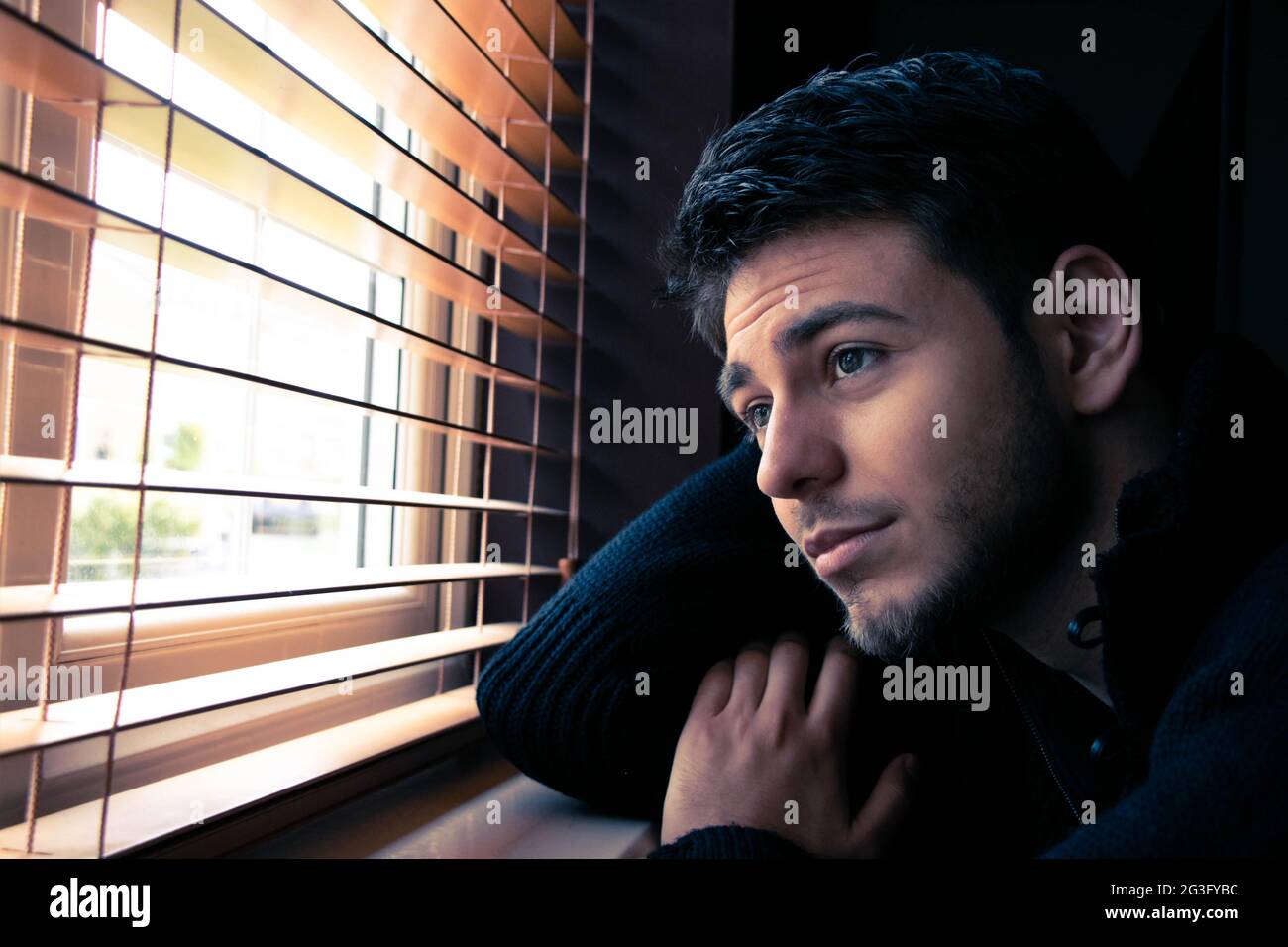 Portrait of attractive man with beard sitting next to window with wood blinds. Stock Photo