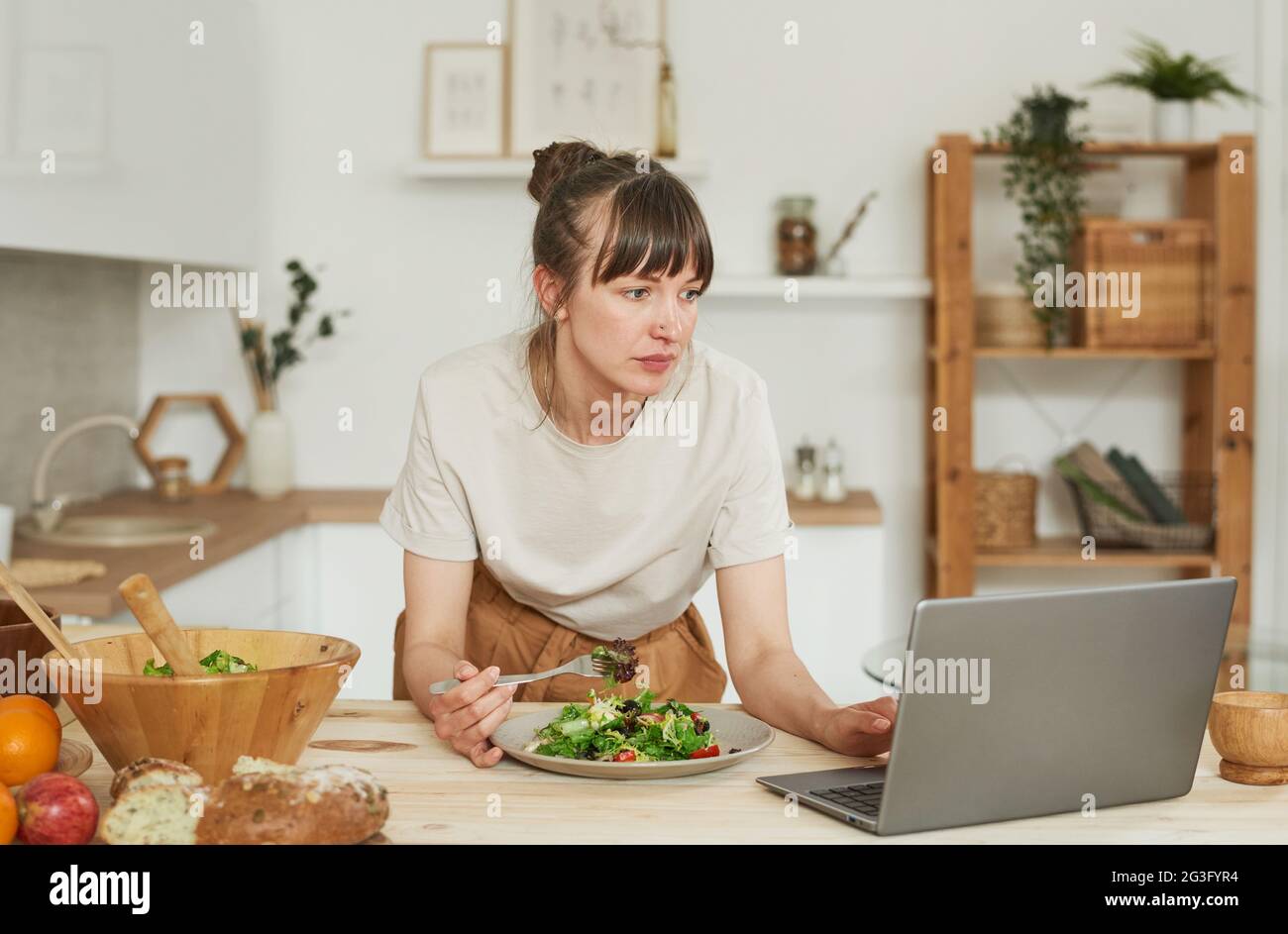 Young woman eating vegetable salad and using laptop in the kitchen Stock Photo