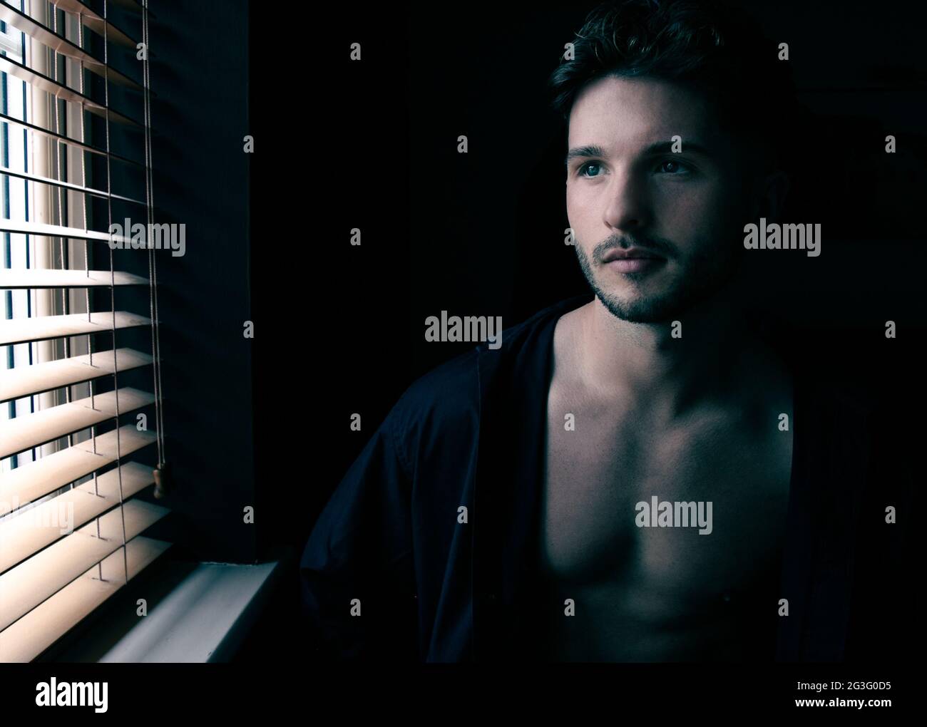 Portrait of attractive shirtless man with stubble sitting next to window with with open shirts and defined pecs Stock Photo
