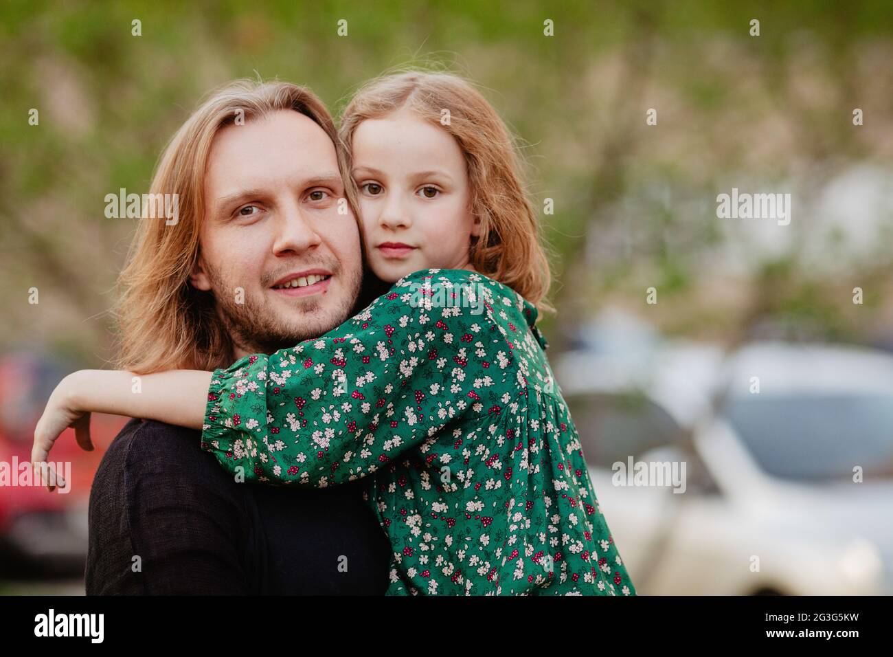 Cute girl gently hugging sliming father in city and looking at camera on blurred background Stock Photo