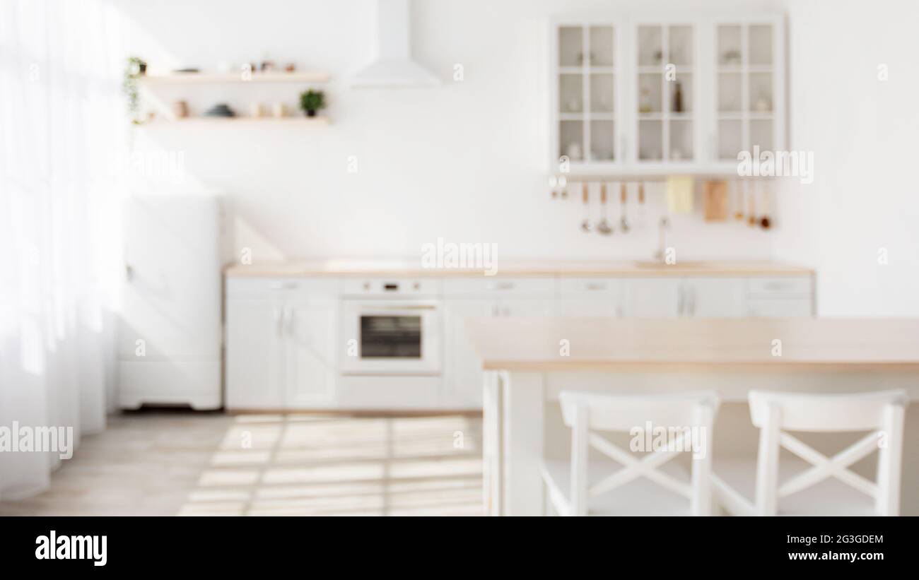 Sunny kitchen interior in Scandinavian style with white kitchen furniture and dining area, blurred background Stock Photo