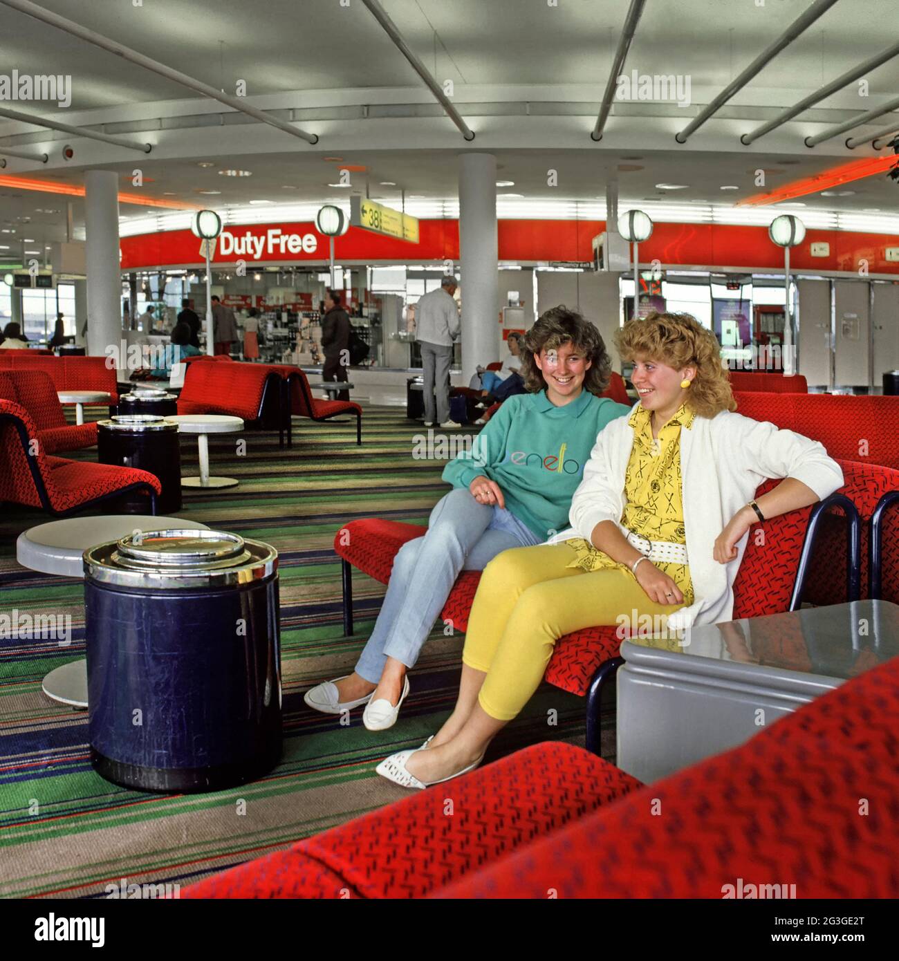 Teenage girls two model released talking and wearing 1980s fashion about to travel to 80s summer family holiday in Spain waiting in the departure lounge building with Duty Free shop & sign beyond at Gatwick Airport West Sussex England UK an historical archive image of the way we were in 1986 Stock Photo