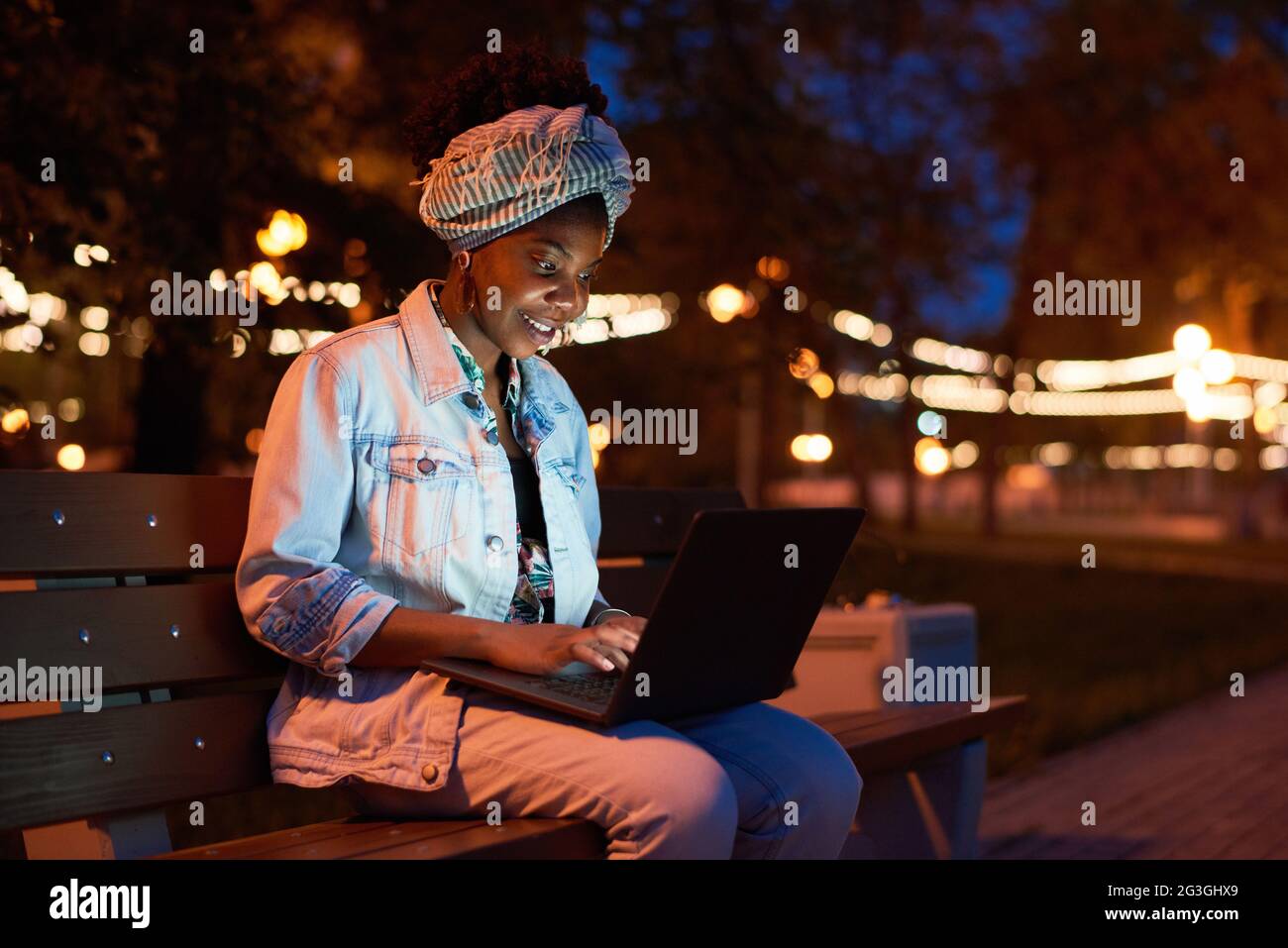 African young woman sitting on bench and using laptop in the park in the evening Stock Photo