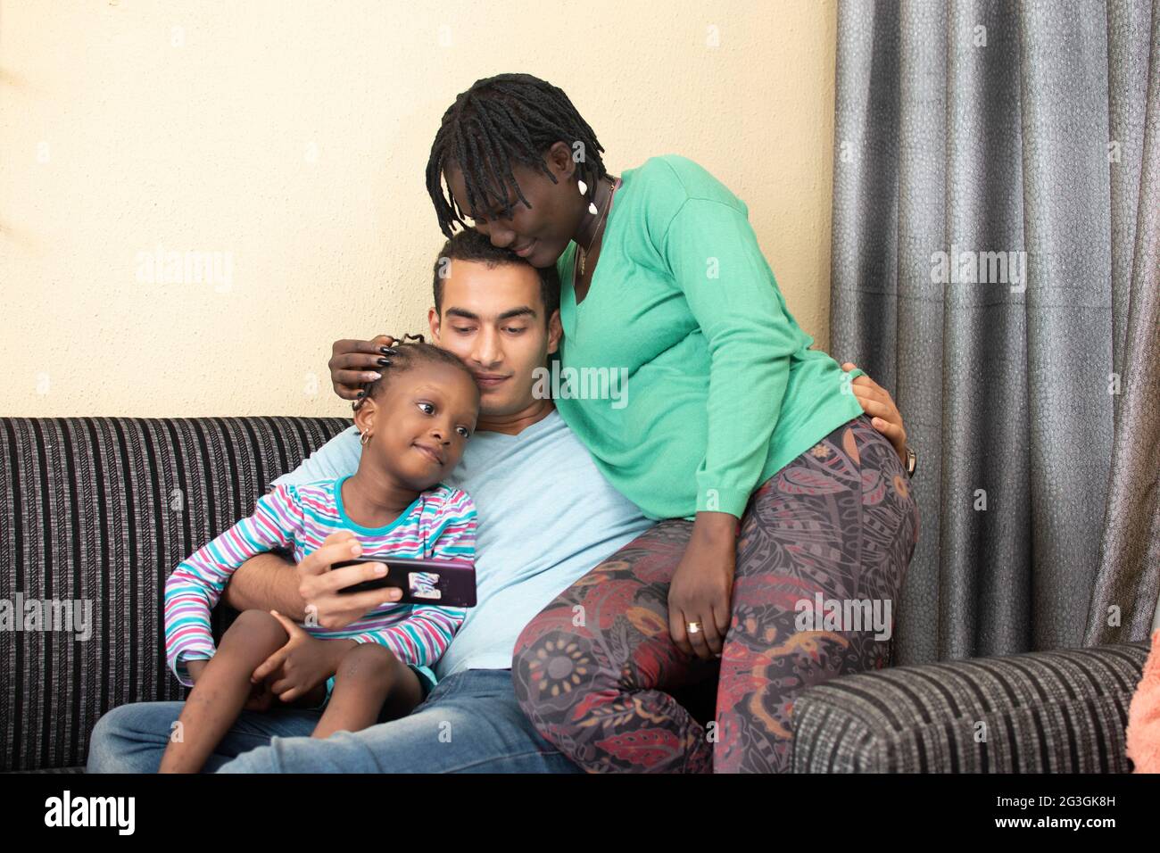 Happy african family mixed race ethnicity parents with small cute little kid child daughter sit on sofa laughing having fun holding smart phone using Stock Photo