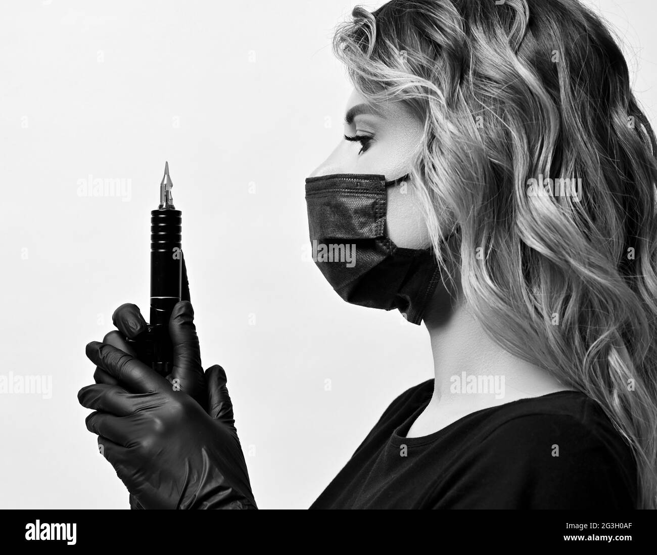Black and white profile of curly woman in black protective medical mask and gloves holding permanent makeup machine Stock Photo
