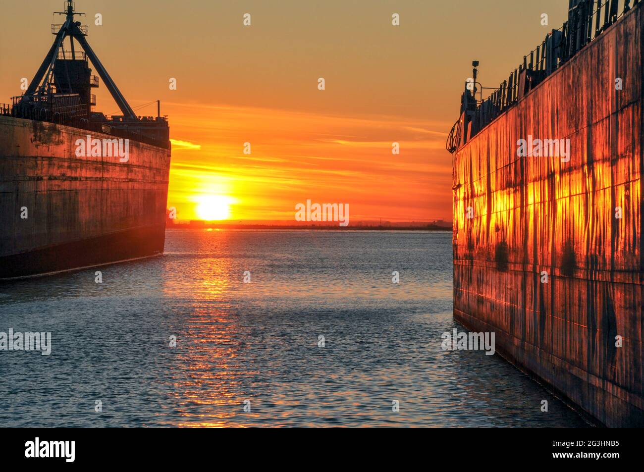 Two bulk carrier laker ships are seen in the port of Toronto, glowing a fiery orange at sunset. Concept of maritime shipping. Stock Photo