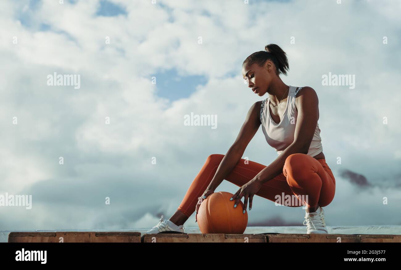 Young woman in sports clothing crouching with basketball outdoors. Sportswoman with medicine ball sitting on rooftop. Stock Photo